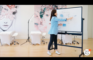 Woman in a blue blazer points at a whiteboard, with artistic wall murals in the background.