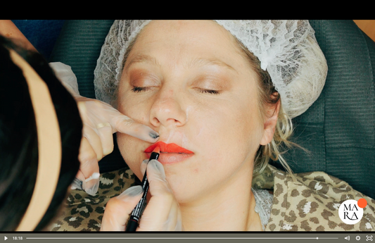 A woman receives lip treatment, eyes closed, with a cosmetics professional applying red lipstick.